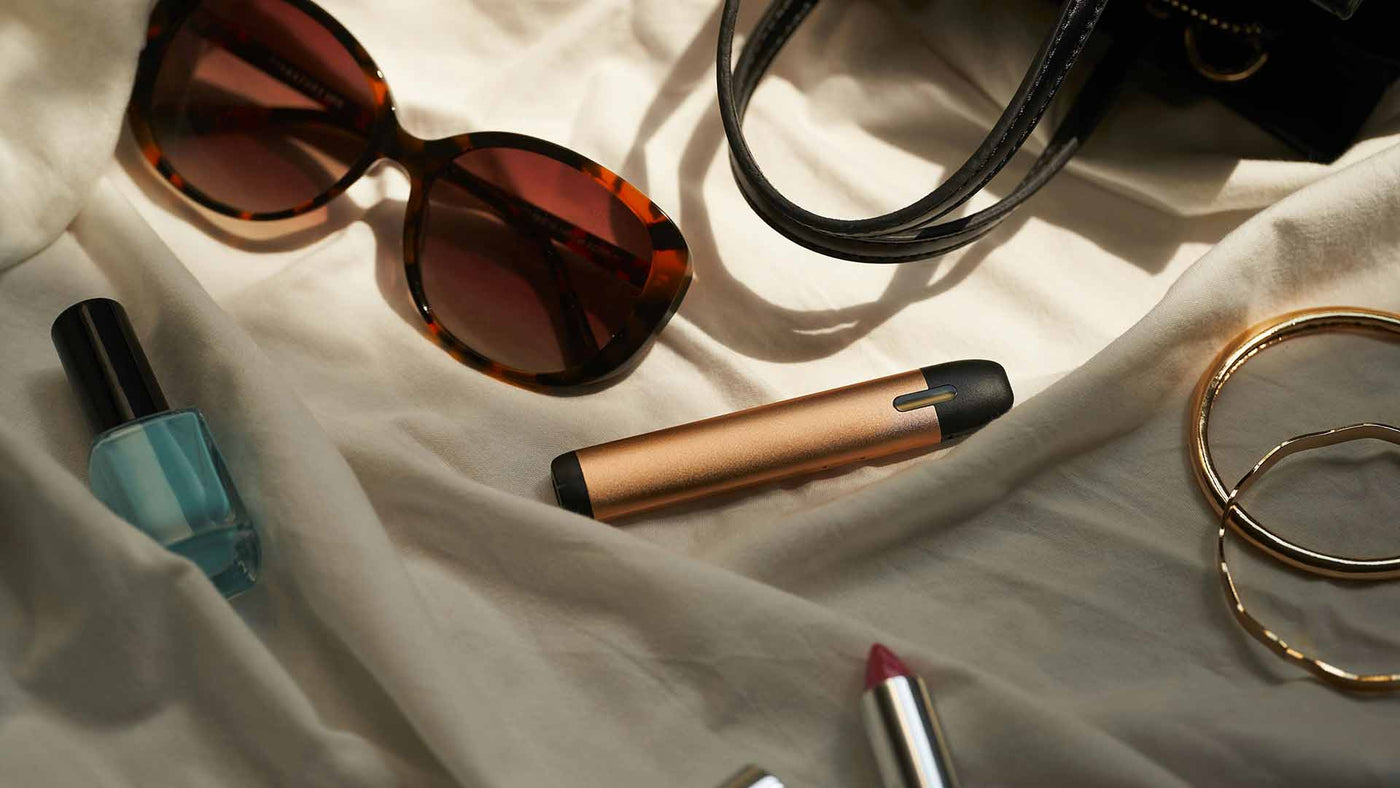 Disposable vape with purse, lipstick, nail polish, sunglasses, and bangles on a bedsheet