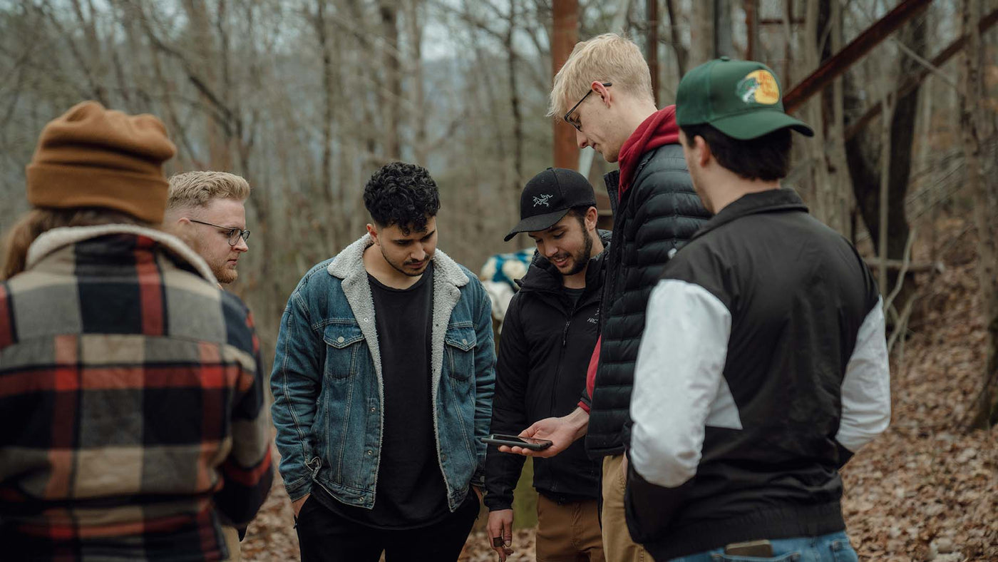 A group of men standing in the woods, looking down at a cellphone.