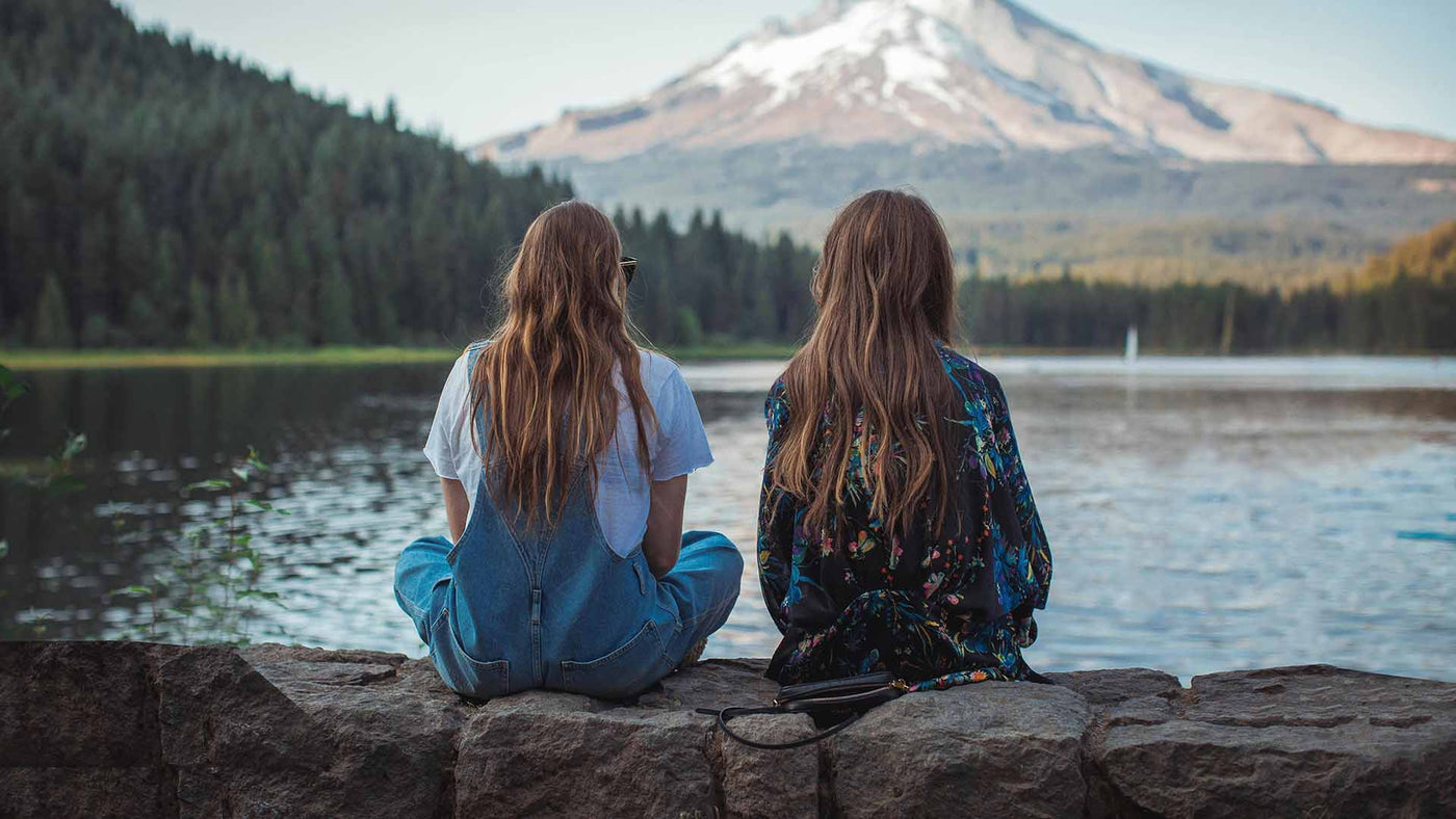 two women sitting on rock facing on body of water and mountain